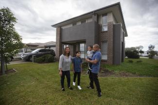 Mother, father and two sons building standing in front of their home smiling